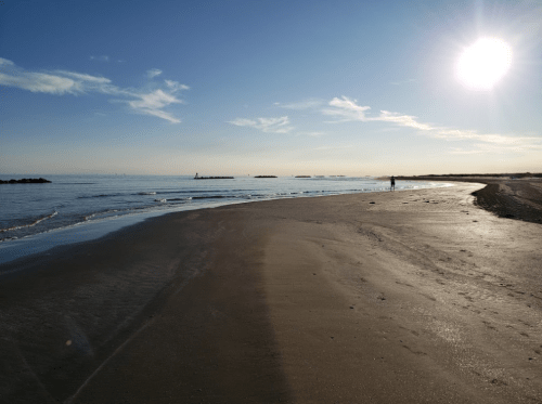 A serene beach scene with gentle waves, a clear sky, and a person walking along the shore under a bright sun.
