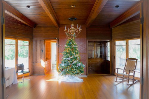 Cozy room with wooden beams, featuring a decorated Christmas tree and a rocking chair, illuminated by natural light.