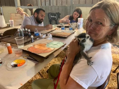 A woman holds a small monkey while others paint at a table in a creative workshop setting.