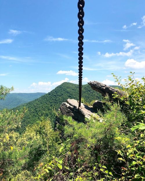 A chain hangs from a rocky outcrop, overlooking lush green mountains under a bright blue sky.