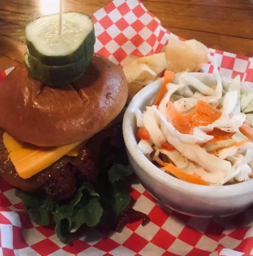 A burger with cheese and pickles on a bun, served with coleslaw and bread rolls on a checkered paper plate.