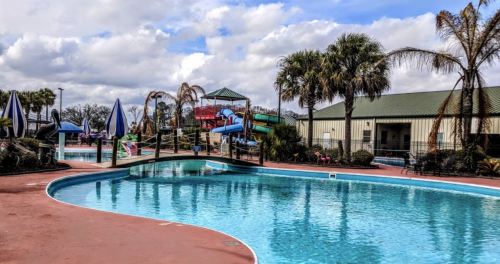 A sunny pool area featuring water slides, palm trees, and lounge chairs under blue skies with scattered clouds.
