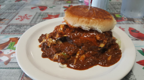 A plate with a sandwich topped with chili, served on a bun, with a festive tablecloth in the background.