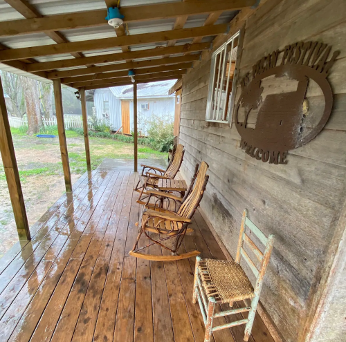 A rustic porch with wooden rocking chairs and a welcome sign, surrounded by greenery and a rainy atmosphere.