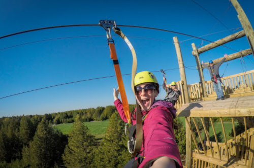 A person in a helmet smiles and poses on a zip line platform, with others zip lining in the background and a green landscape below.