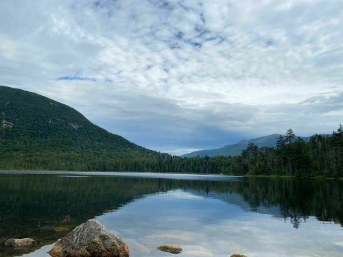 A serene lake surrounded by mountains and lush trees, reflecting the cloudy sky above.