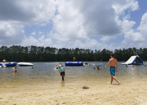 A sandy beach with people swimming and playing on inflatable structures in a lake under a partly cloudy sky.