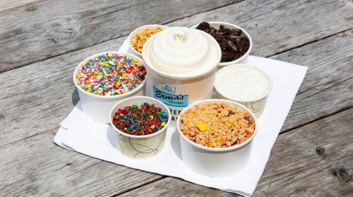 A variety of ice cream toppings and a tub of soft serve on a wooden surface, arranged on a white napkin.