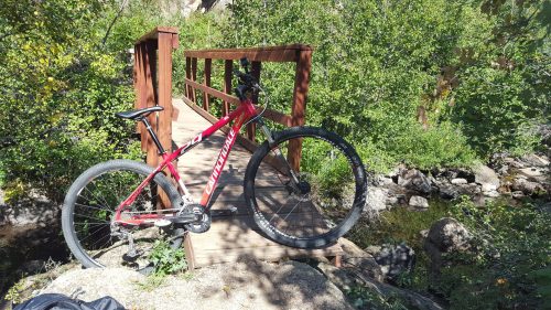 A red mountain bike rests on a wooden bridge surrounded by greenery and rocks near a stream.