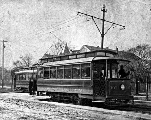 A vintage streetcar with passengers, stopped on a dirt road, surrounded by trees and utility poles.