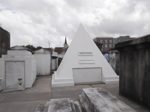 A white pyramid-shaped tomb in a cemetery, surrounded by stone crypts and cloudy skies.