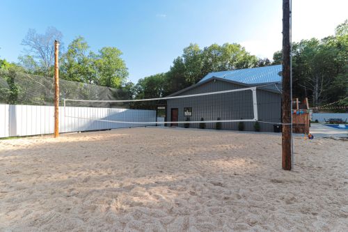 A sandy volleyball court with a net, surrounded by trees and a building in the background.