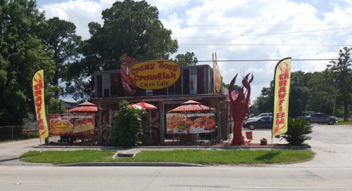A colorful Cajun café with a sign reading "Crawfish" and vibrant flags outside, surrounded by greenery and blue skies.