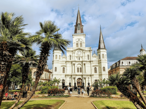 A historic church with tall spires surrounded by palm trees and people walking in a vibrant park setting.