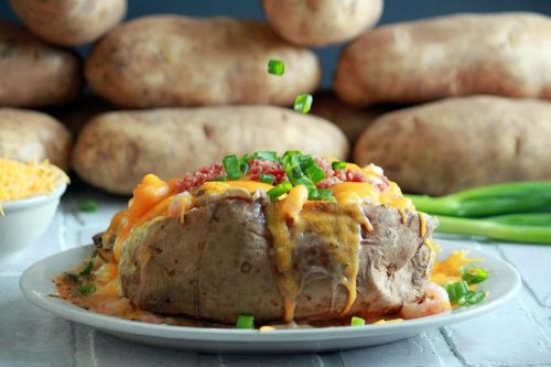 A loaded baked potato topped with cheese, bacon, and green onions, with whole potatoes in the background.
