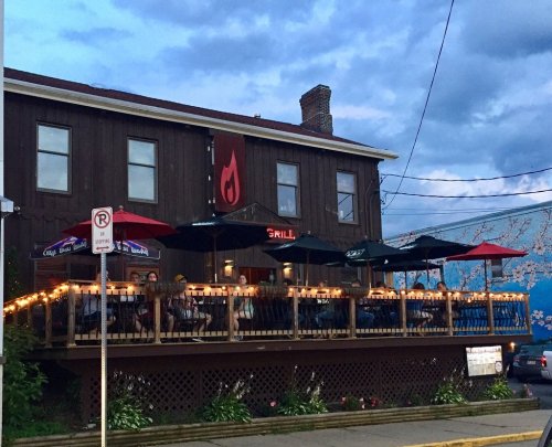 A cozy restaurant with outdoor seating, string lights, and umbrellas, set against a cloudy sky.