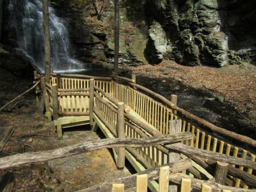 A wooden walkway with railings leads to a waterfall, surrounded by rocky cliffs and autumn leaves.