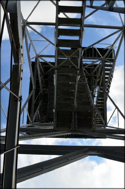 View from below a metal structure, showcasing stairs and beams against a blue sky with clouds.