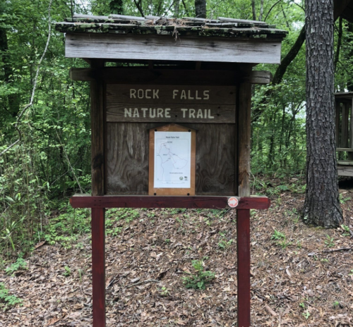 Wooden sign for Rock Falls Nature Trail, featuring a map and surrounded by lush greenery.