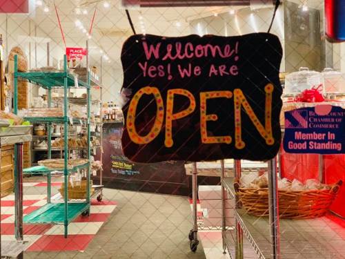 A colorful sign in a shop window reads "Welcome! Yes! We Are OPEN," with shelves and baked goods visible behind it.