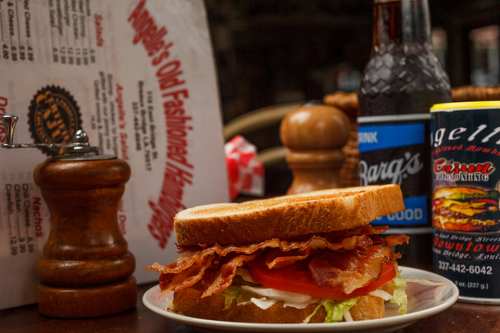 A close-up of a sandwich with bacon, lettuce, and tomato, alongside a soda and a menu in a diner setting.