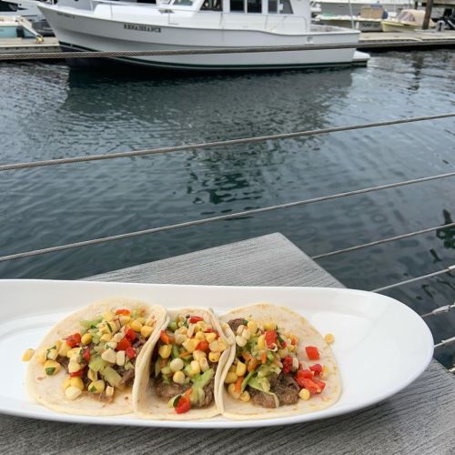 Three tacos filled with meat, corn, and vegetables on a plate, with a marina and boat in the background.