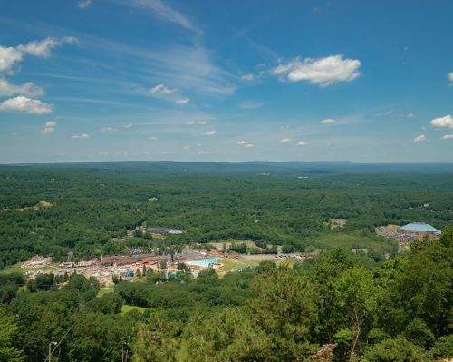 A panoramic view of lush green hills and a distant town under a bright blue sky with scattered clouds.