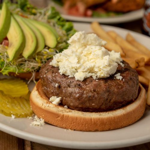 A juicy burger topped with crumbled cheese on a bun, served with avocado, salad, fries, and pickles on the side.