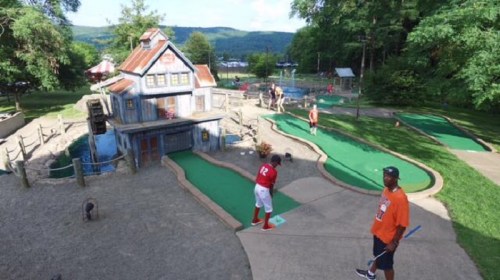 A mini-golf course with a whimsical building, featuring players on the green and trees in the background.