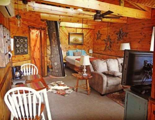 Cozy cabin interior with wooden walls, a sofa, dining area, and a bed in a rustic setting.