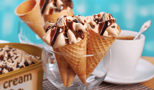 Three ice cream cones topped with chocolate sauce and whipped cream, with a bowl of ice cream and a cup in the background.