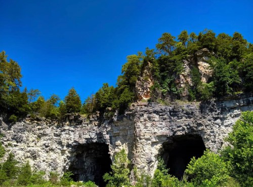 A rocky cliff with two large openings, surrounded by lush green trees under a clear blue sky.