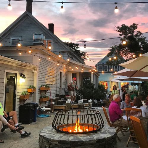 A cozy outdoor gathering with string lights, a fire pit, and people enjoying music and conversation at sunset.