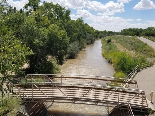 A wooden bridge spans a muddy river surrounded by lush greenery under a partly cloudy sky.