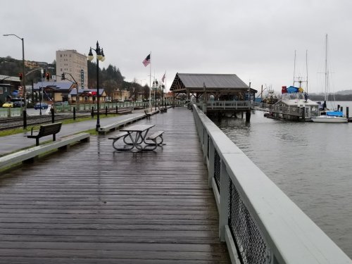 A rainy waterfront pier with benches, a gazebo, and boats in the water, surrounded by buildings and flags.