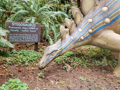 A Stegosaurus statue with a sign explaining its name and characteristics, surrounded by greenery.
