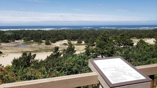 A scenic view of the ocean and forest from an elevated viewpoint, with a map sign in the foreground.