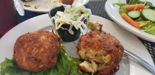 Two golden-brown crab cakes on lettuce, served with a side of coleslaw and a fresh garden salad.