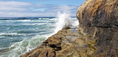 Waves crashing against rocky shore under a cloudy sky, with ocean water splashing onto the rocks.