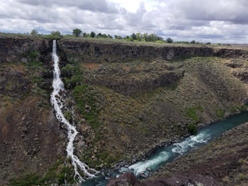 A waterfall cascades down a rocky cliff into a river below, surrounded by rugged terrain and cloudy skies.