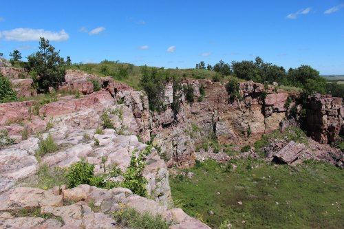 A rocky cliff with colorful layers, surrounded by greenery and a clear blue sky.