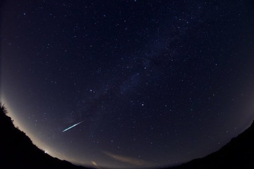 A starry night sky with a bright meteor streaking across the Milky Way, framed by dark silhouettes of mountains.