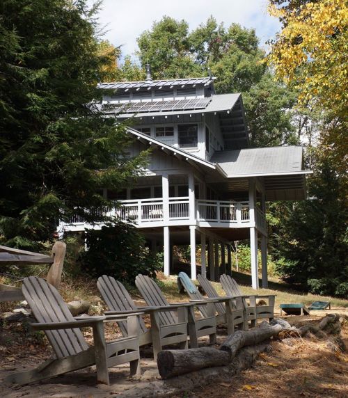 A modern gray house on stilts surrounded by trees, with wooden chairs arranged in front on a grassy area.