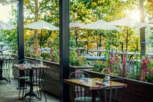 Outdoor restaurant patio with tables, chairs, and vibrant flowers, surrounded by trees and umbrellas in the background.