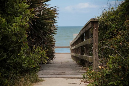 A wooden path leads through lush greenery to the ocean, under a partly cloudy sky.