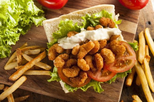 A sandwich with crispy shrimp, lettuce, tomatoes, and mayonnaise, served with fries on a wooden board.
