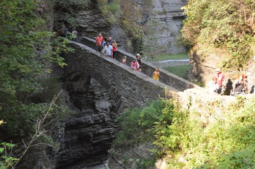 A stone bridge spans a rocky gorge, with people walking along it surrounded by lush greenery and steep cliffs.