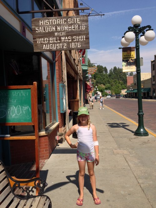 A child stands on a sidewalk in front of a historic saloon sign, wearing a green cap and summer attire.