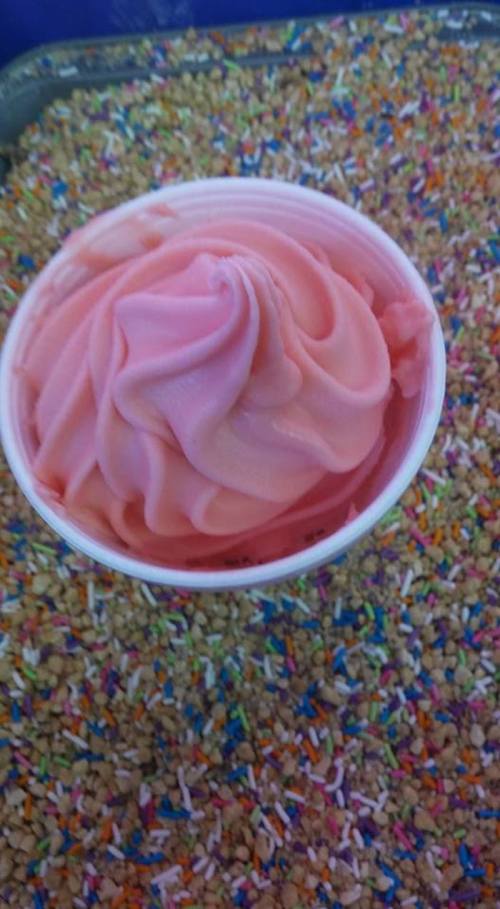 A cup of pink soft serve ice cream topped with colorful sprinkles, resting on a bed of sprinkles.