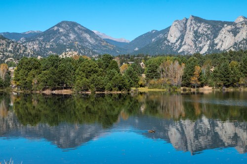 A serene lake reflects mountains and trees under a clear blue sky, showcasing vibrant autumn colors.